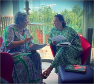 2 women talking sitting in red chairs, both holding papers and a book.